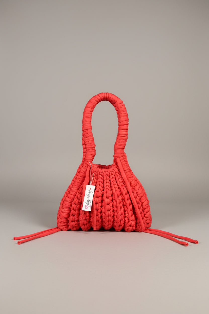 A bright red hand-woven macrame shoulder bag with a single integrated handle, laid flat on a neutral gray background. A white paper tag with the brand name 'Jenapara' is attached to the side.