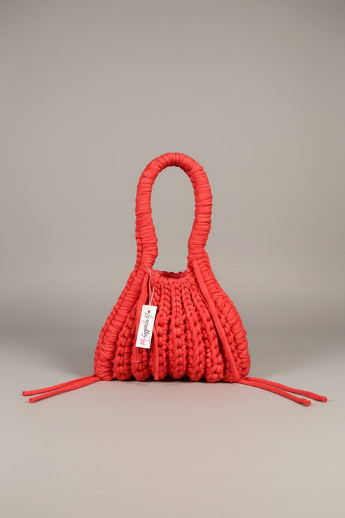 A bright red hand-woven macrame shoulder bag with a single integrated handle, laid flat on a neutral gray background. A white paper tag with the brand name 'Jenapara' is attached to the side.