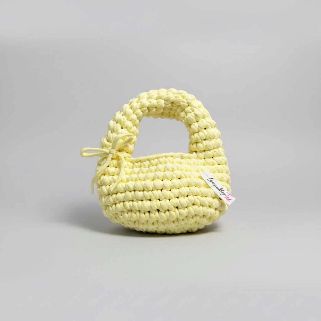 A pale yellow crocheted baby bib with a small bow on the left side and a small white brand tag, displayed on a beige fabric background.